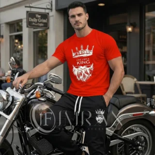 A male fashion model, for 'IF Imran Fabric ,' is shown posing on a vintage motorcycle parked on a city street outside a cafe with 'Daily Grind' signage. He is wearing the crimson red t-shirt and black joggers set featuring the white crown and 'Live Like a King" text and Beard graphic.