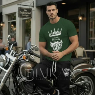 A male fashion model, for 'IF Imran Fabric ,' is shown posing on a vintage motorcycle parked on a city street outside a cafe with 'Daily Grind' signage. He is wearing the hunter green t-shirt and black joggers set featuring the white crown and 'Live Like a King" text and Beard graphic.