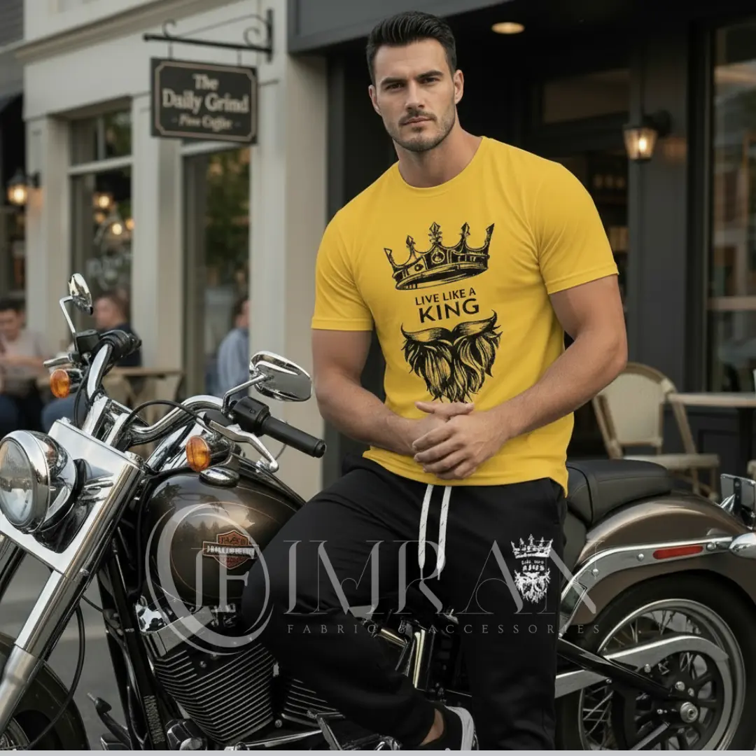 A male fashion model, for 'IF Imran Fabric ,' is shown posing on a vintage motorcycle parked on a city street outside a cafe with 'Daily Grind' signage. He is wearing the sun-kissed Gold t-shirt and black joggers set featuring the white crown and 'Live Like a King" text and Beard graphic.