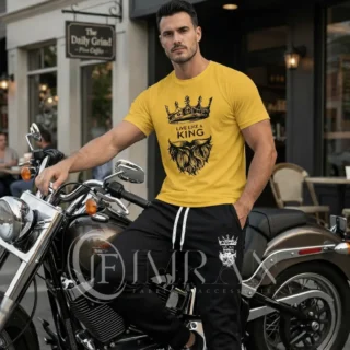 A male fashion model, for 'IF Imran Fabric ,' is shown posing on a vintage motorcycle parked on a city street outside a cafe with 'Daily Grind' signage. He is wearing the sun-kissed Gold t-shirt and black joggers set featuring the white crown and 'Live Like a King" text and Beard graphic.
